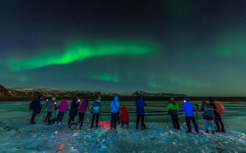 A large group of aurora watchers with tripods lined up on an icy shore under green northern lights