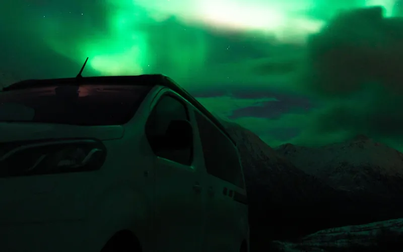 White minibus parked below snowy mountains near Tromsø under a bright green aurora