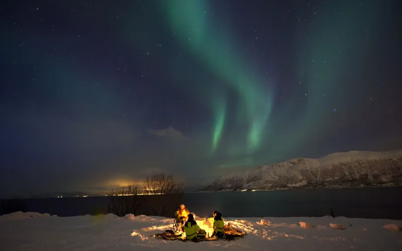 A few people around a campfire on a snowy fjord shore near Tromsø under the northern lights